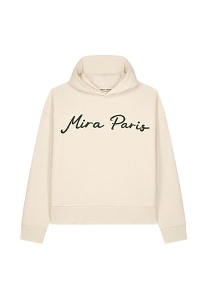 Donna con capelli biondi lisci indossa occhiali da sole neri e una felpa beige con testo "Mira Paris", abbinata a pantaloni scuri su sfondo bianco.