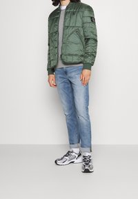 Zielona pikowana kurtka bombka z nylonowego materiału, z prążkowanymi mankietami i dołem. Pod spodem szara koszulka z okrągłym dekoltem, zestawiona z jasnoniebieskimi dżinsami i trampkami.