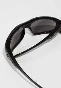 Óculos de sol pretos com uma estrutura de plástico elegante, lentes escuras e detalhes em dourado da marca. O design é desportivo com uma forma envolvente.