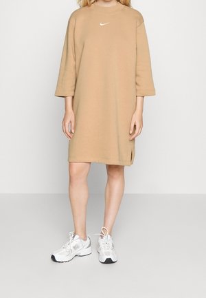 Kobieta w beżowej, oversize sukience typu sweatshirt marki Nike z rękawami do łokcia oraz białych butach sportowych New Balance stoi na szarej podłodze.