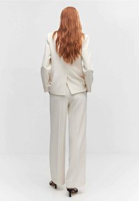 Costume couleur crème avec un blazer structuré et un pantalon à jambes larges. Tissu lisse, design sur mesure, et une fente centrale au dos. Escarpins noirs.