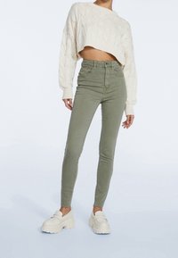 Suéter recortado de punto trenzado color crema, jeans ajustados de talle alto en verde olivo y mocasines de plataforma blancos. Textura suave con un diseño ajustado.