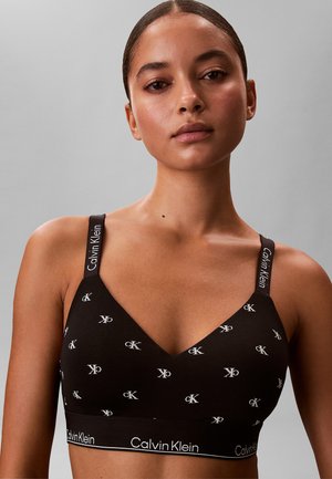 Kvinde iført sort Calvin Klein bralette med hvidt logo og mærkede stropper, mod en ensfarvet grå baggrund.
