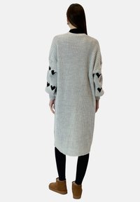 Jasnoszary sweter kardigan w czarne wzory serc na rękawach, o długim kroju, oversize, z fakturowanej tkaniny, w zestawie z czarnymi legginsami i brązowymi butami.