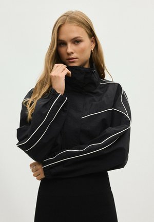 Jeune femme aux longs cheveux blonds portant une veste noire avec un passepoil blanc, tenant le col d'une main sur un fond uni.