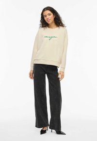 Beige genser med grønn kursiv brodering av "imagine", kombinert med høytlivs sorte vide bukser og svarte spisse hæler.