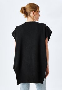 Chaleco negro de punto oversize con mangas cortas, que presenta un corte holgado y aberturas laterales, llevado sobre unos jeans de un azul claro. Textura suave.
