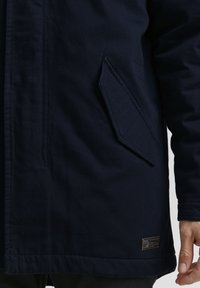 Navy blue outerwear jacket featuring a textured fabric, patch pocket, and a small metal logo tag at the hem.