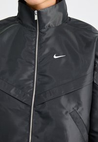 Svart Nike-jacka med hög krage, zip-up fram och vit logotyp. Tillverkad av ett slätt, texturerat tyg med modern design och vinklade sömmar.