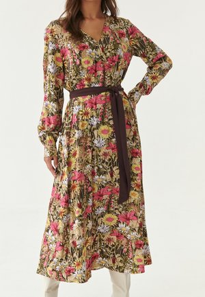 Mujer vestida con un vestido midi de manga larga con estampado floral multicolor y cinturón oscuro en la cintura, combinado con botas de color claro, posando con una mano en la cadera.