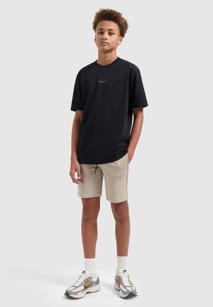 Tienerjongen staat met één hand in de zak van beige shorts, draagt een zwart T-shirt, witte sokken en witte met gouden sneakers op een grijze achtergrond.