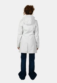 Impermeabile bianco con cappuccio, caratterizzato da una vestibilità su misura e una texture liscia. Abbinato a jeans a zampa d'elefante blu scuro, visto da dietro.