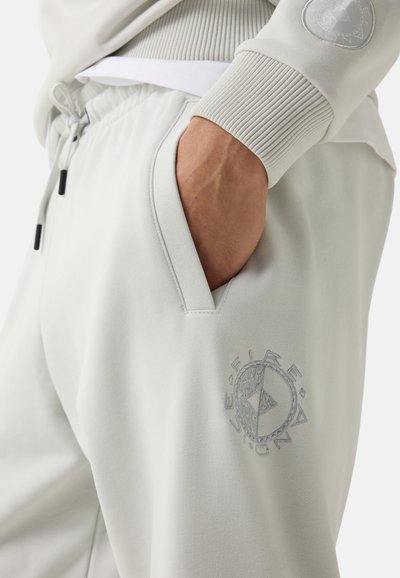 Close-up van een persoon die lichtgrijze joggingbroek draagt met een hand in de zak en een cirkelvormig embleem op de dij, gecombineerd met een bijpassend sweatshirt.