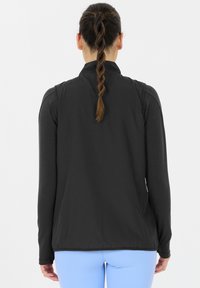 Svart sportväst med långa ärmar, slät textur, dragkedja fram och en figurnära design, kombinerad med ljusblå byxor.