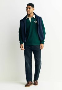 Tume roheline pika varrukaga polosärk beeži kraega ja tumesinine kampsun õlgadel; koos tumedate teksade ja pruunide kingadega.