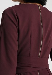 Robe bordeaux à manches longues, avec une fermeture éclair dorée visible dans le dos et une ceinture en tissu pour une silhouette ajustée. Texture douce.