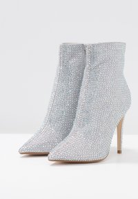 Stříbrné kotníkové boty pokryté průhlednými rhinestony, špičaté špičky, vysoký stiletto podpatek, hladká textura a zip na zadní straně pro zapínání.
