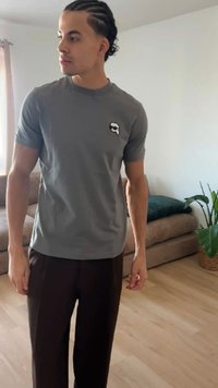 Hombre con el pelo trenzado, de pie en el interior, lleva una camiseta gris con un pequeño logo y pantalones oscuros, cerca de un sofá y una planta en maceta.