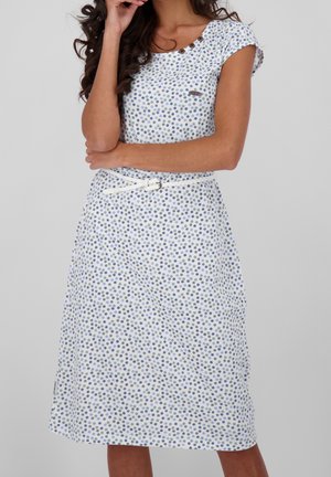 Kvinde iført hvid kjole med blå og grå prikker, korte ærmer og et hvidt bælte, stående foran en ensfarvet baggrund.