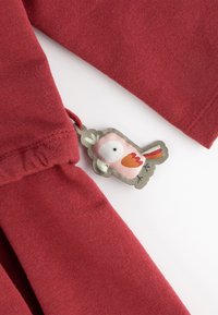 Roter Baumwollstoff mit gerafftem Ärmel und einem Stoffetikett mit einem pinken Vogel-Motiv, akzentuiert mit orangefarbenen, grünen und schwarzen Details.