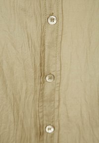 Sección de camisa de tela arrugada beige que presenta cinco botones circulares de color claro a lo largo de la tapeta delantera y costuras cosidas.