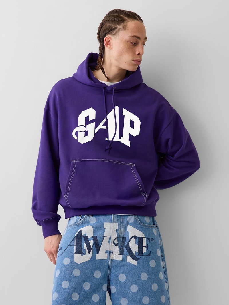 Personne aux cheveux tressés portant un sweat à capuche violet GAP et un jean bleu à pois avec "AWAKE" imprimé sur le devant.
