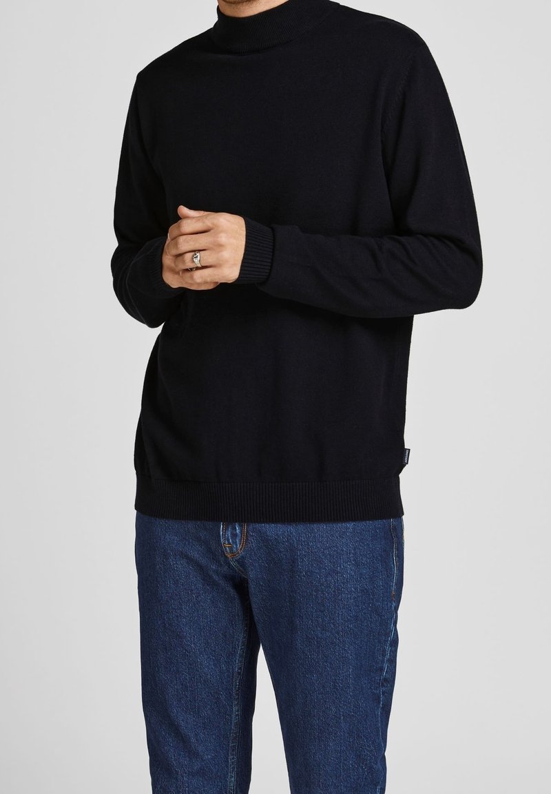 Pull noir à col roulé tricoté avec poignets et ourlet côtelés, porté par-dessus un jean bleu. Conception ajustée avec une texture lisse.