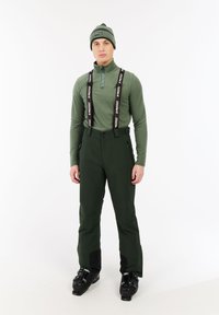 Man in groene wintersportkleding, waaronder een beanie, een ritsjack, hoge broek met bretels en zwarte skiboots op een witte achtergrond.