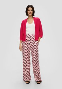 Rosa geripptes Cardigan, weißes enges Oberteil und weite Hosen mit einem Herzmuster in Rot und Dunkelrosa auf einem weißen Hintergrund. Schwarze Sandalen.