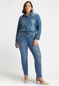 Koszula dżinsowa z guzikiem z przodu, dwoma kieszeniami na piersi i akcentami w postaci łańcucha; połączona z dopasowanymi, niebieskimi dżinsami i jasnymi, spiczastymi obcasami.