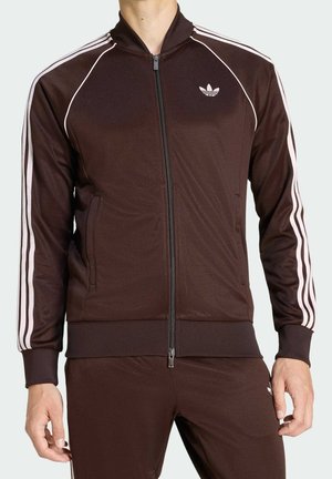 Mann trägt braunen Adidas-Trainingsanzug mit weißen Streifen an Ärmeln und Hosen, steht mit entspannten Armen an den Seiten.