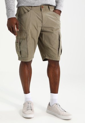 Dunkelhäutige Person trägt beige Cargo-Shorts, weiße Socken, hellbeige Sneaker und einen grauen Pullover, steht mit den Händen in den Taschen.