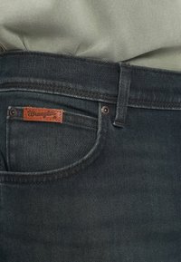Tamsiai pilki džinsai, su artimu kišenės vaizdu ir rudos odos Wrangler etikete. Ypatybės: užtrauktukas ir mygtuko užsegimas.