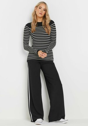 Langsleeve zwart-wit gestreepte top, gecombineerd met losse zwarte broek met witte zijstrepen en witte sneakers. Gladde stof textuur.