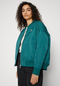 Teal bomberjakke med lynlås foran, ribbede manchetter og et subtilt logo på brystet. Lavet af glat, letvægtsmateriale.