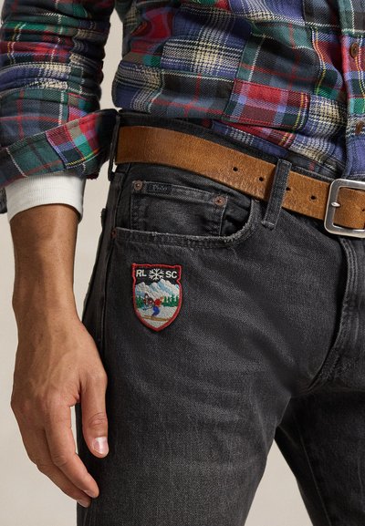 Μαύρο τζιν από τζιν υλικό με ένα πολύχρωμο κεντητό μπαλώμα που απεικονίζει ένα τοπίο, συνδυασμένο με μια καφέ δερμάτινη ζώνη και ένα καρό πουκάμισο.