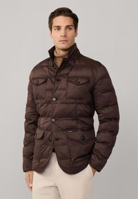 Chaqueta acolchada marrón con cierre de botones, dos bolsillos en el pecho y dos bolsillos inferiores, con cuello alto y textura acolchada.