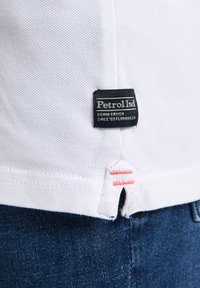 Valkoinen polo-paita, jossa on teksturoitu kangas, musta "Petrol Ind" -tunnus ja punaiset yksityiskohdat alareunassa tummansinisten farkkujen päällä.