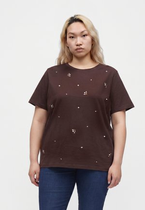Kvinde med langt blondt hår iført en brun t-shirt dekoreret med små blomsterprint og blå jeans, stående mod en ensfarvet baggrund.