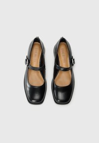Pair of black leather Mary Jane shoes with square toes and ankle straps, beige insoles branded "Steve Madden," on a plain gray background.