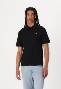 Svart polo skjorte med korte ermer, ribbestrikket krage og en liten hvit logo på brystet, kombinert med lyseblå denimjeans.