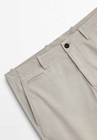 Pantalon beige ajusté avec passants de ceinture, poches à l'italienne sur les côtés et à l'arrière, et fermeture à bouton noir sur la ceinture.