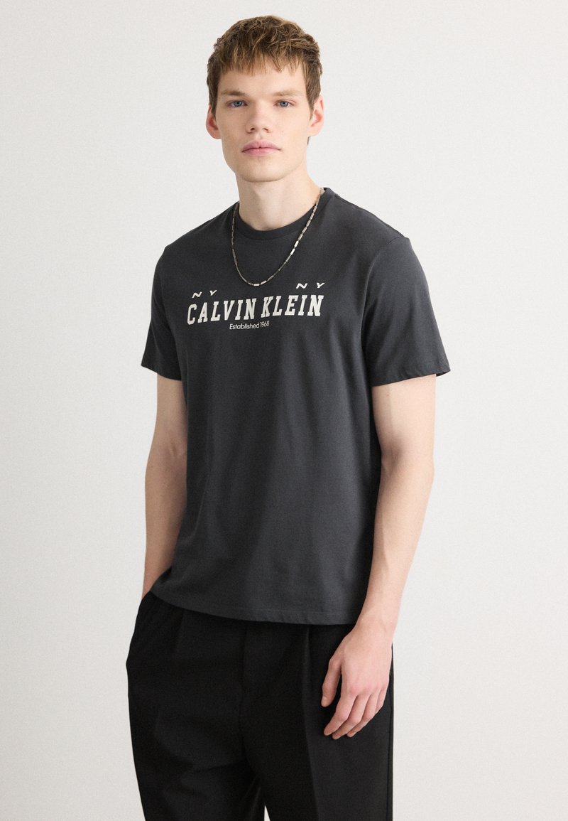 Must küllastatud puuvillane T-särk, millel on valge "CALVIN KLEIN" logo, ümmargune kaelus, lühikesed varrukad ja lõdva istumisega. Sobib vabade mustade pükstega.