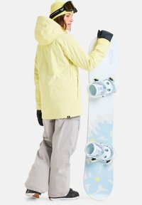 Snowboard met lichtblauw bloemenpatroon, witte bindingen en een gele quilted jas over grijze sneeuwbroek. Zwarte handschoenen en een skibril.