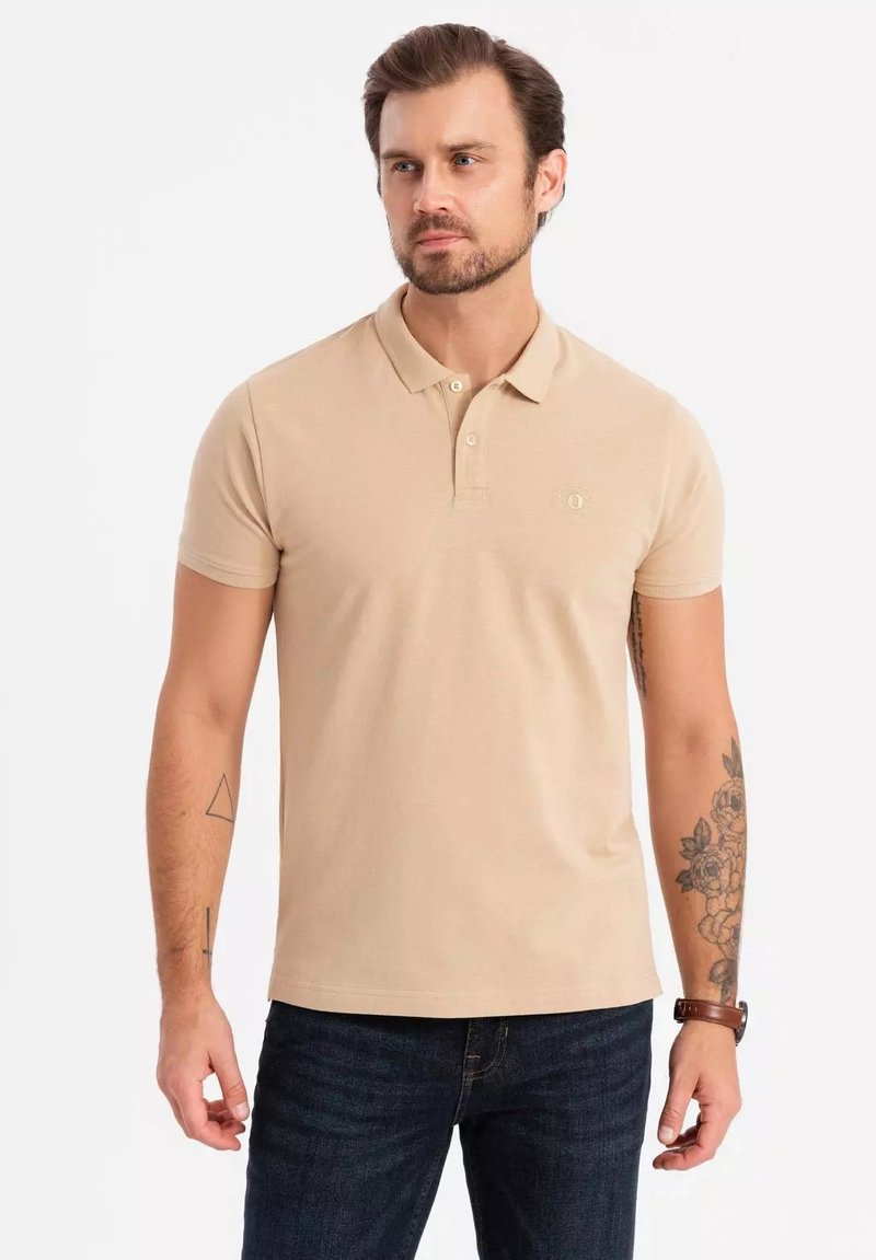 Beige polo tričko vyrobené z texturované látky, s límečkem na tři knoflíky a malým logem na hrudi. Nosené s tmavými džínami.