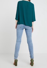 Niebieska bluzka z rękawami o długości trzech czwartych, luźny krój, zestawiona z jasnoniebieskimi obcisłymi dżinsami i zielonymi butami na słupku. Prosty i nowoczesny design.