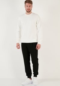 Felpa bianca a maniche lunghe con scollo rotondo, abbinata a pantaloni jogger neri. Il modello indossa sneakers bianche. Design semplice e casual.