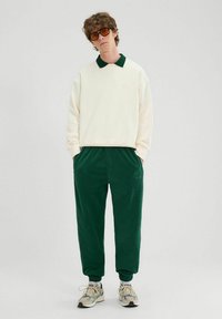 Kremowa bluza z zielonym kołnierzykiem, w połączeniu z ciemnozielonymi dżinsami dresowymi. Zauważalne struktury w materiale; sneakersy mają wielokolorowy wzór.