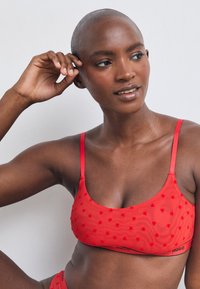 Rød bralette med hjerte mønster, tynne stropper og en logo-bånd. Laget av myk stoff, med en glatt tekstur og tettsittende design.