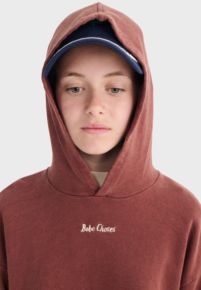 Junge Person trägt eine rötlich-braune Kapuzenjacke mit dem Aufschrift "Bobo Choses" und eine marineblaue Kappe, schaut nach unten mit einem neutralen Gesichtsausdruck.